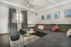 A modern living room with a grey sectional sofa, a glass coffee table, and a round ottoman. There are three framed artworks on the wall and a window with grey curtains.