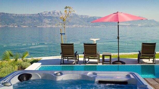 A poolside area with a hot tub, lounge chairs, and a table with an umbrella, overlooking a body of water and mountains. - Beachfront Villa Grey Martini with Pool (Split)