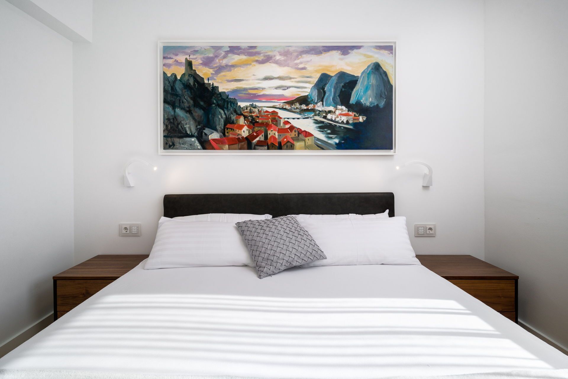 A neatly made bed with white linens and a gray pillow, a dark headboard, and a painting of a coastal town above it.