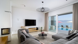 A modern living room with a flat-screen TV, grey sofas, a small coffee table, and a view of a pool and sea through large sliding glass doors. - Beachfront Villa Grey Martini with Pool (Split)