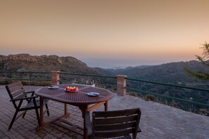 Ein Tisch mit Stühlen auf einer terrassenförmig angelegten Pflasterterrasse mit Blick auf eine gebirgige Landschaft.