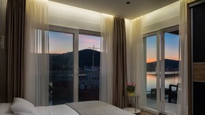 A hotel room with a large bed, a small table with a vase, and a balcony with a view of the water and cityscape. - Beachfront Villa Seashell with Pool (Marina)