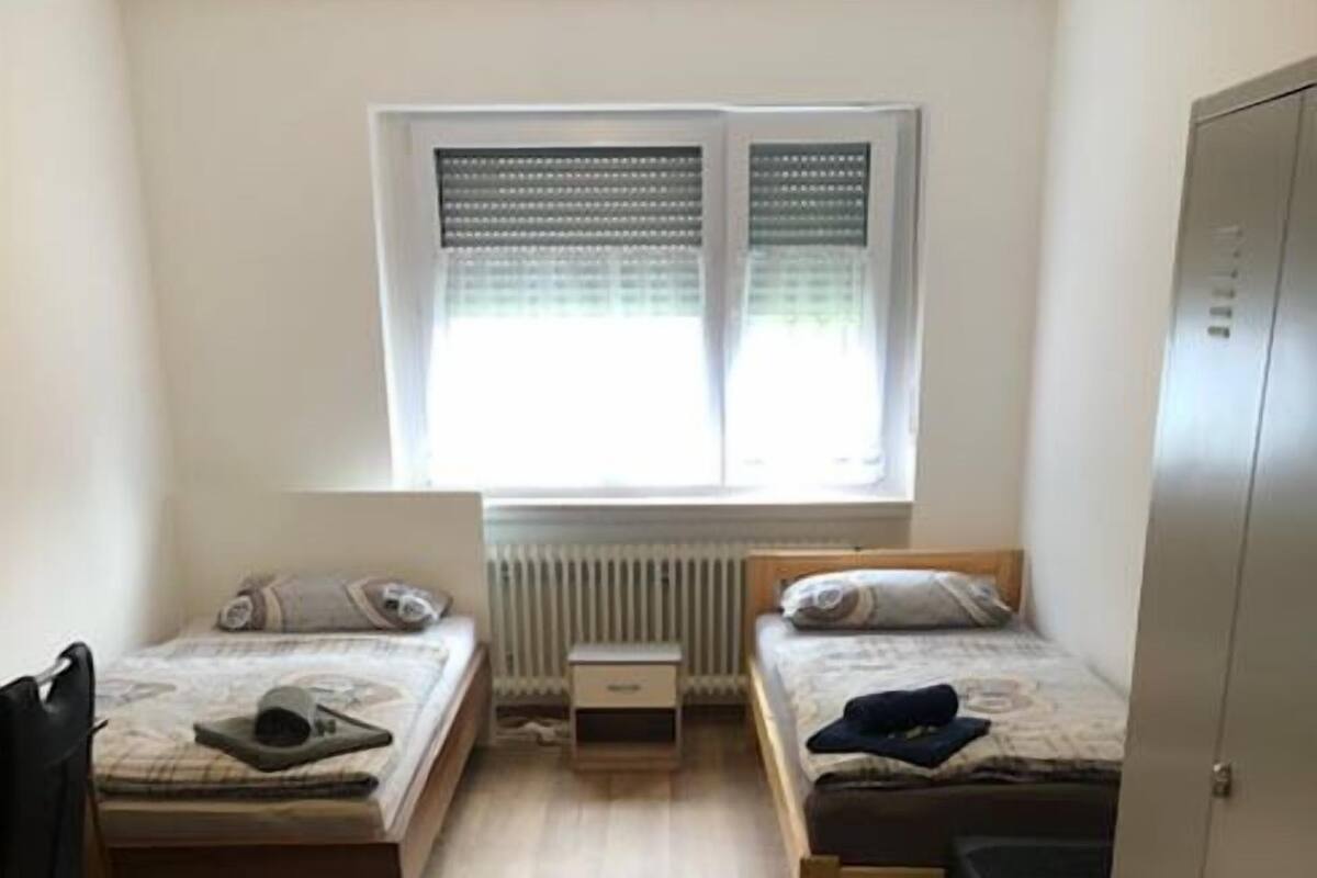 Een compacte, goed verlichte kamer met twee éénpersoonsbedden, een radiator, een raam met luxaflex en een kledingkast.