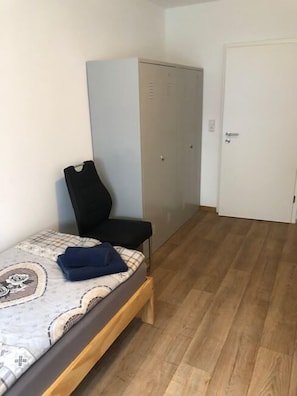 Ein schlichter, spärlich eingerichteter Raum mit einem Bett, einem Stuhl und einem Kleiderschrank.