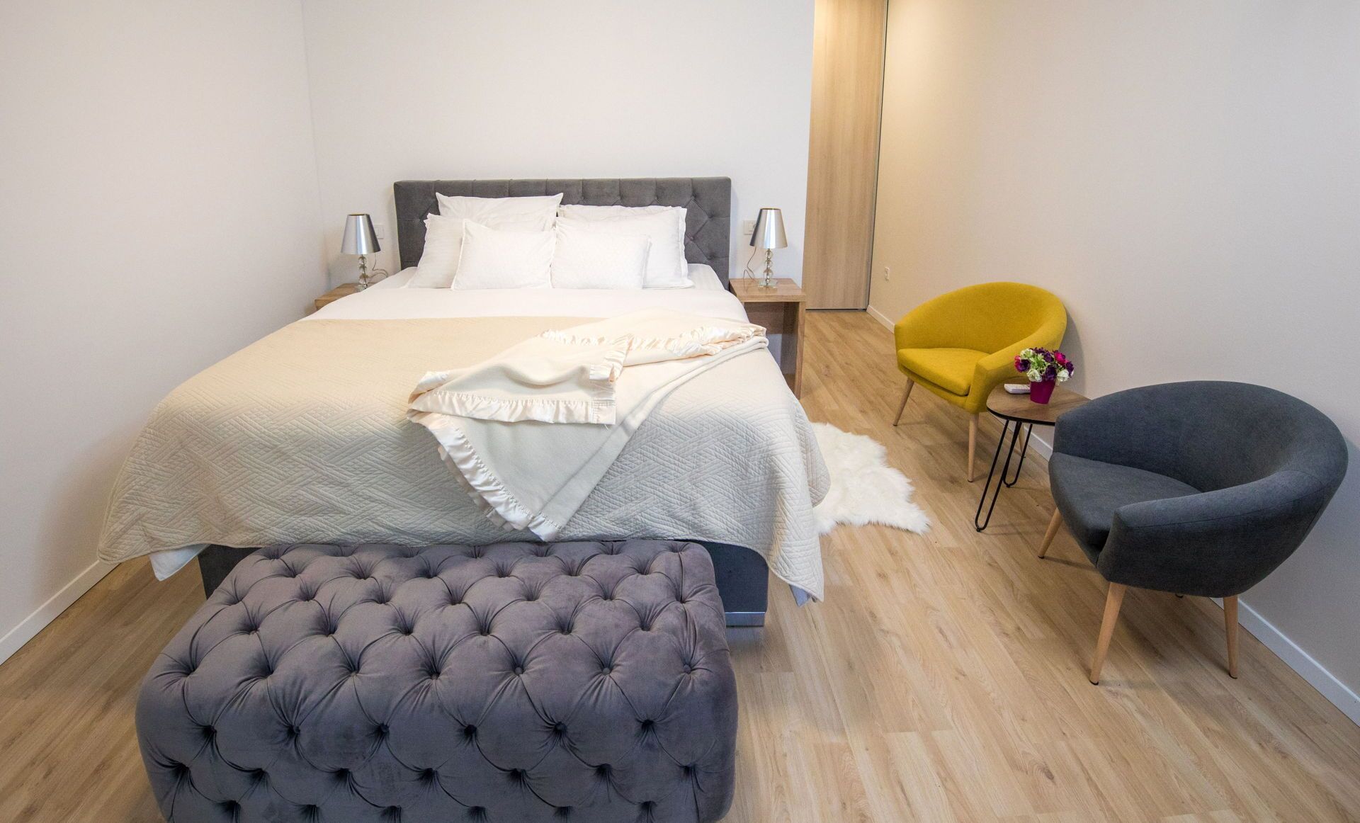 A neatly arranged bedroom with a bed, bedside lamps, a small table, a chair, and a tufted ottoman.