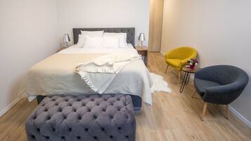 A neatly arranged bedroom with a bed, bedside lamps, a small table, a chair, and a tufted ottoman.