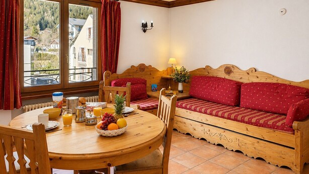 A cozy room with a wooden dining table, chairs, a sofa, and a view of a mountainous landscape through a window. - Cozy apartment in Chamonix center with parking (Chamonix-Mont-Blanc)