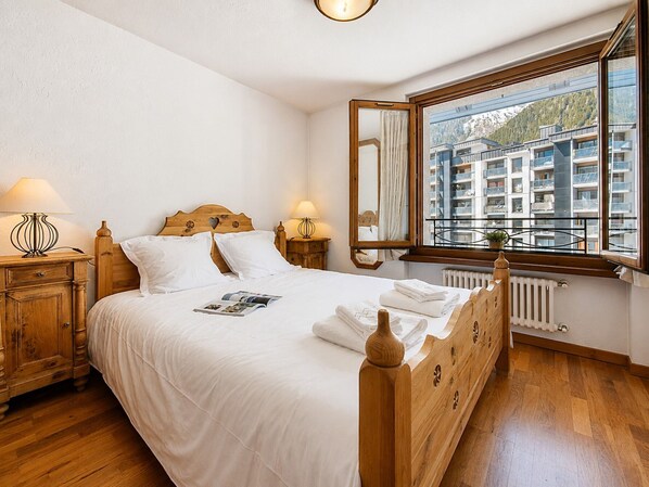 A neatly made bed with a wooden headboard, a bedside table with a lamp, a window with a view of buildings, and a radiator.