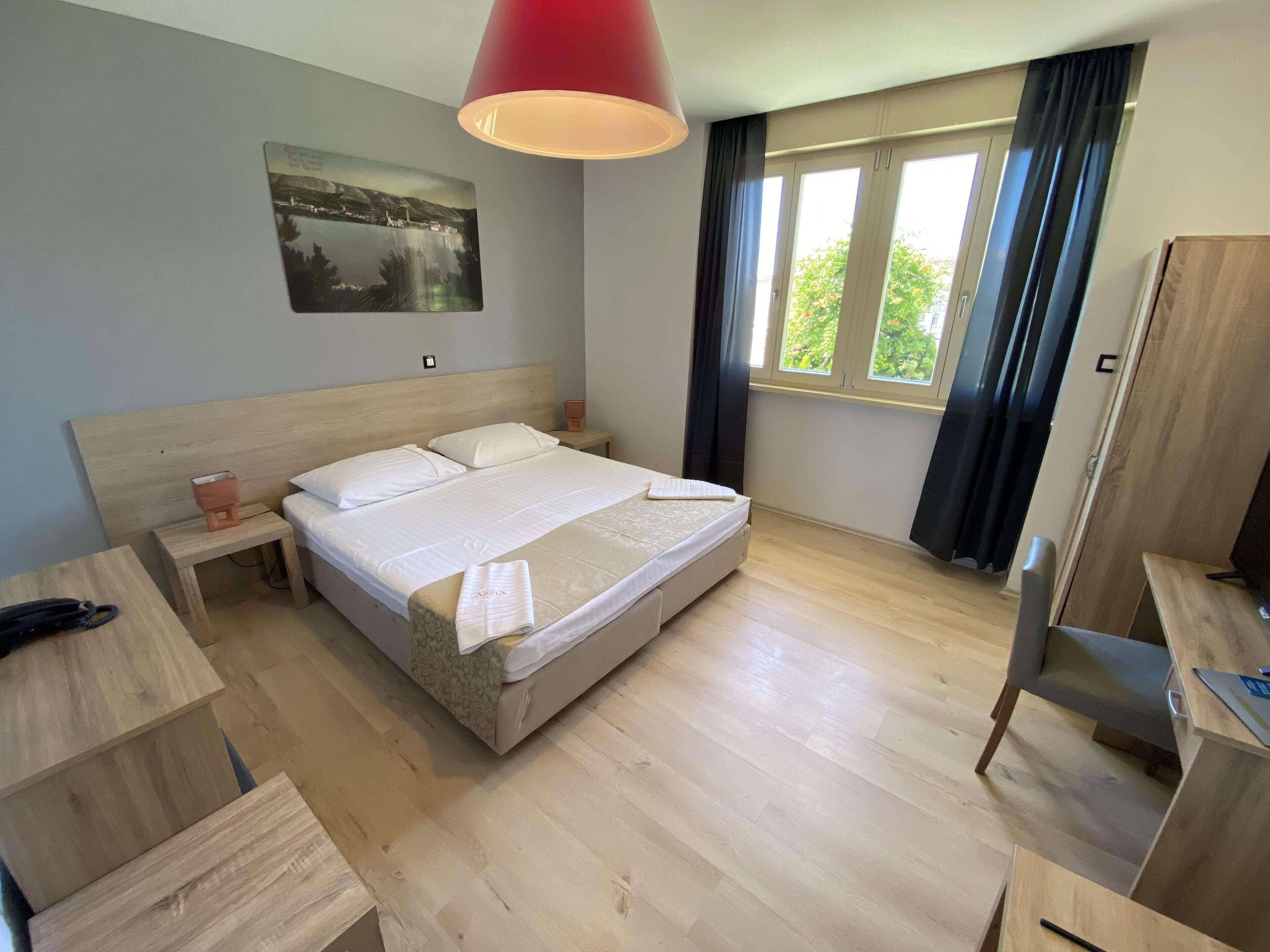 Ein modernes Hotelzimmer mit einem hölzernen Bett, einem Schreibtisch, einem Stuhl, einer roten Lampe, einem Fenster mit Vorhängen und einem Bild an der Wand.