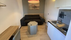 Un espacio habitable compacto con kitchenette, un sofá gris, una mesa de madera y una lámpara colgante roja.