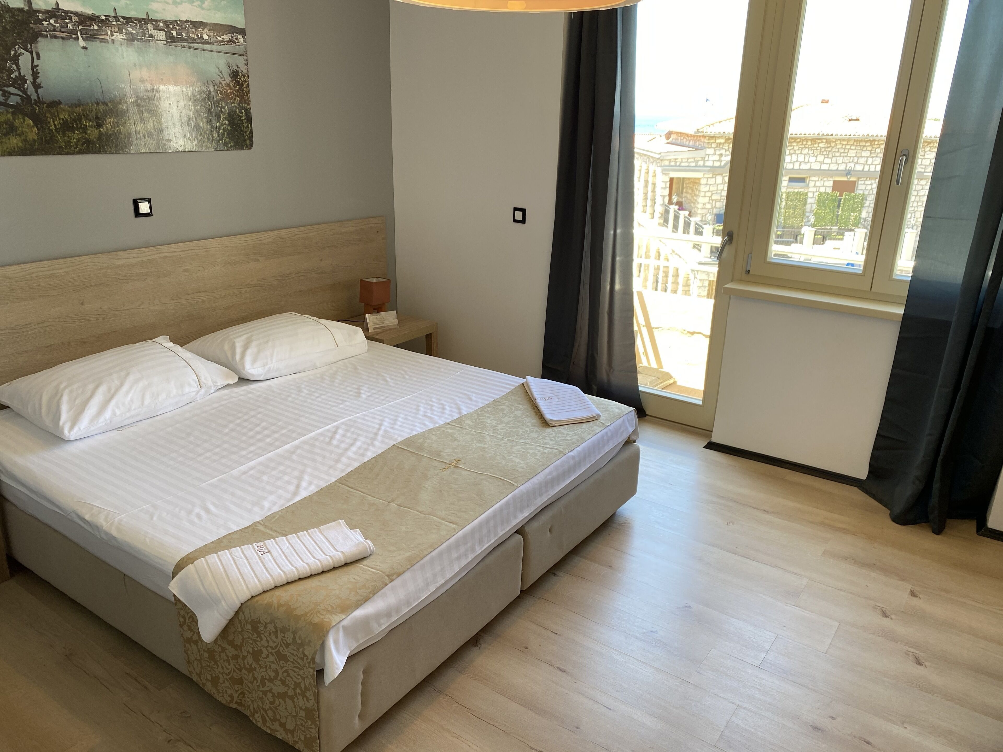 A neatly made bed with white linens and a beige throw blanket, a wooden headboard, a bedside table with a lamp, and a view of a building through a window with curtains.