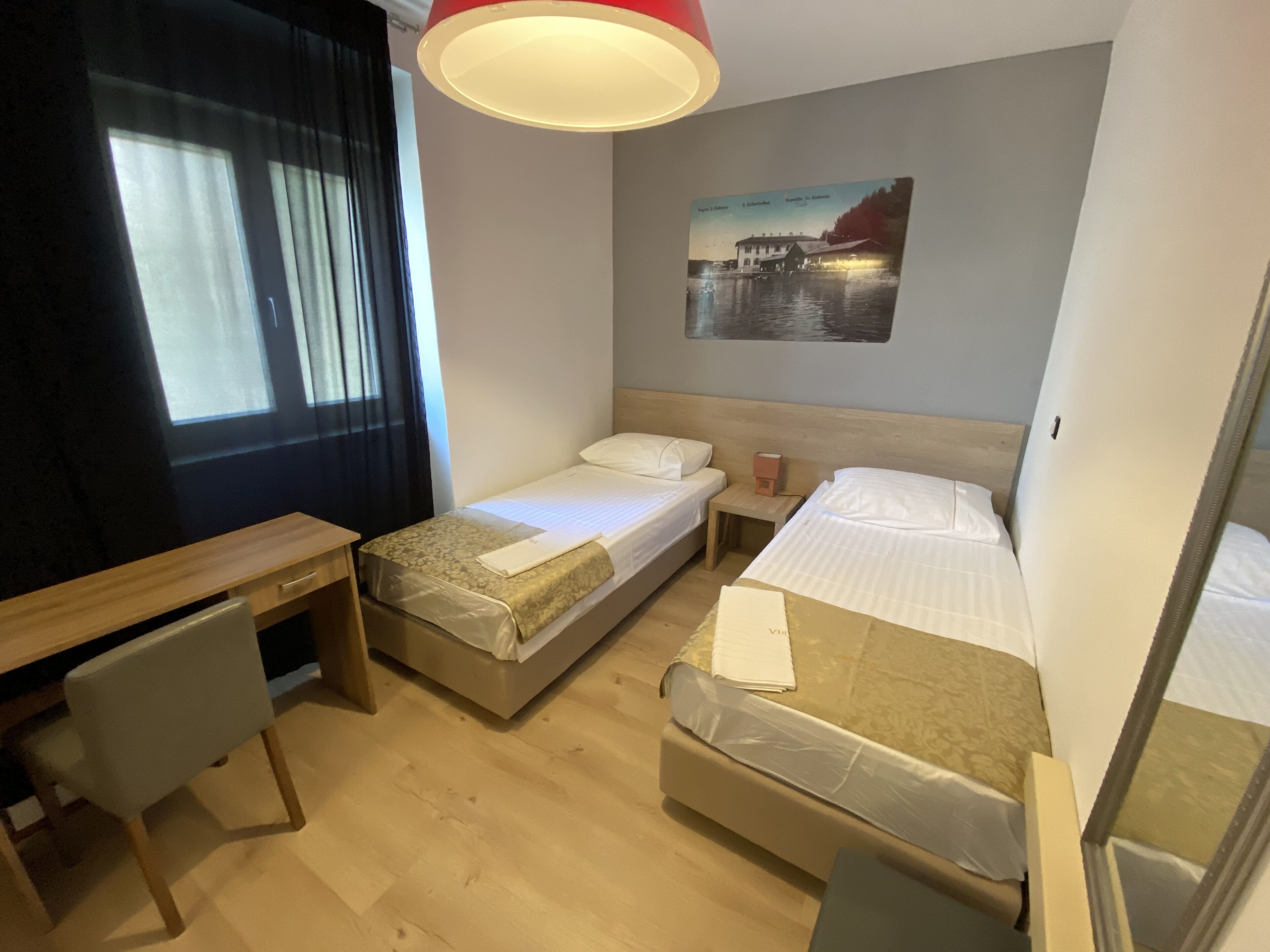 A hotel room with two single beds, a desk, a chair, a small table, and a wall-mounted picture.
