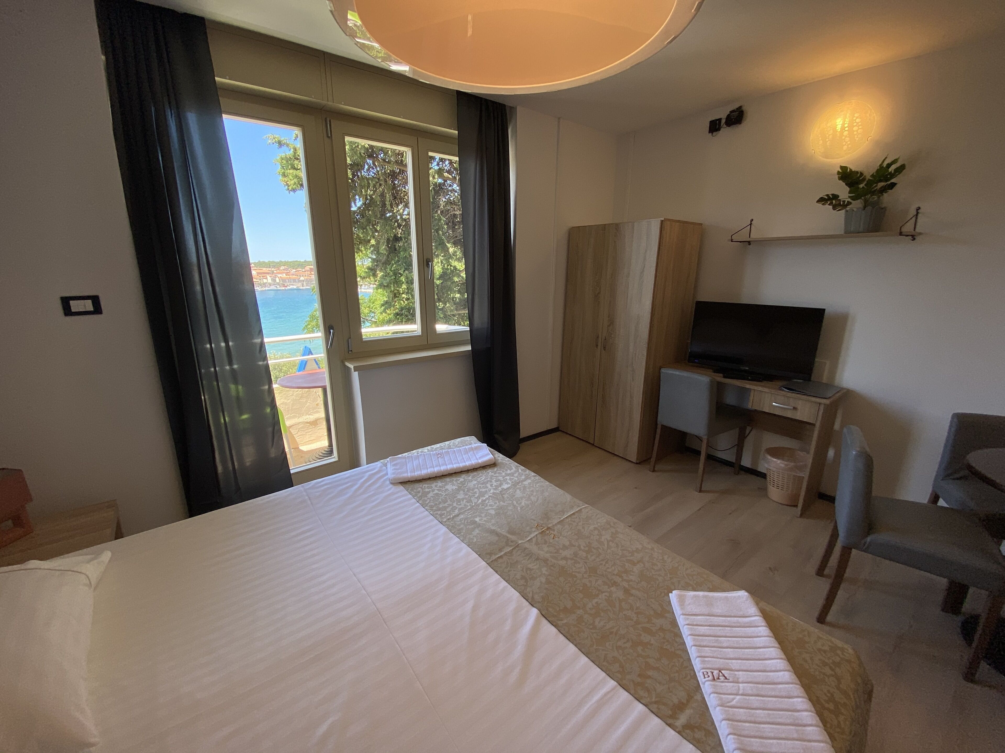 A hotel room with a bed, a desk with a chair, a TV, and a view of the sea through large windows.