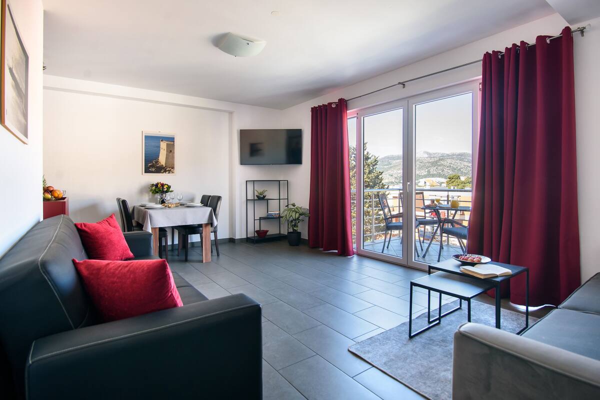 Un salon moderne avec un canapé en cuir noir, des coussins rouges, une table à manger avec des chaises, un téléviseur à écran plat et un balcon agrémenté de rideaux rouges.