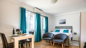 A modern bedroom with a bed, two chairs, a dining table, and a window with curtains. - California Apartments II (Dubrovnik)
