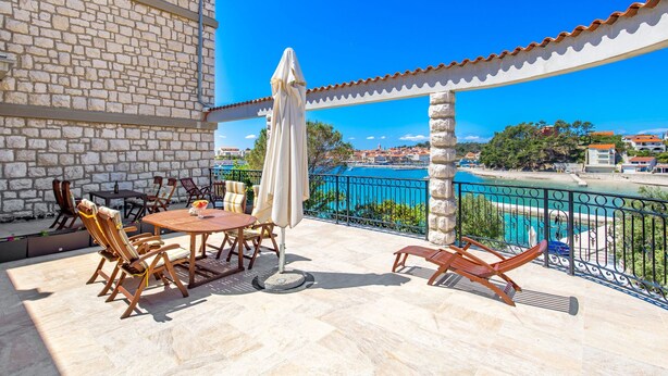 Une terrasse avec des murs en pierre, une table et des chaises, un parasol, et une vue sur une mer bleue et des bâtiments.