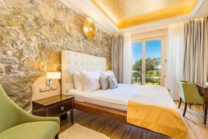 A hotel room with a stone wall, a large bed with a white headboard, a nightstand with a lamp, a desk, and a view of the outdoors. - Integrated Hotel Arbia - Margita Deluxe (Rab)