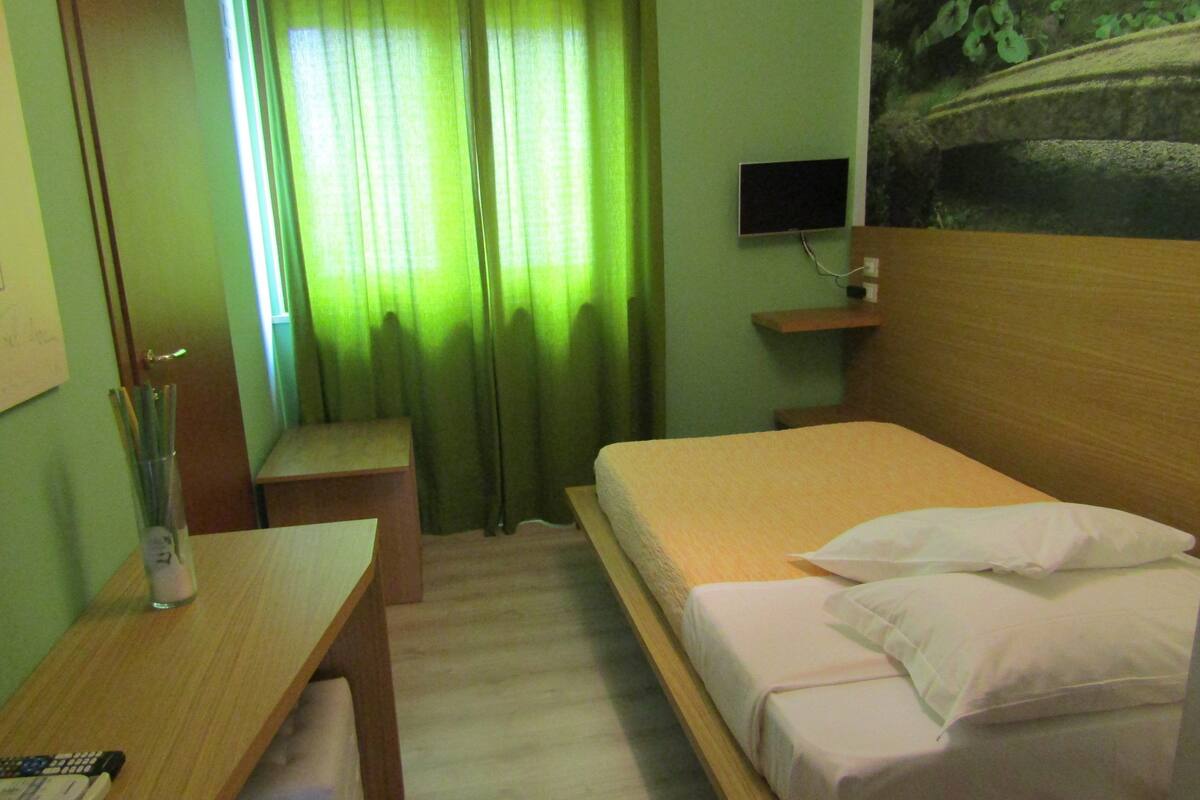 A hotel room with a bed, a desk, a chair, a TV, and a window with curtains.