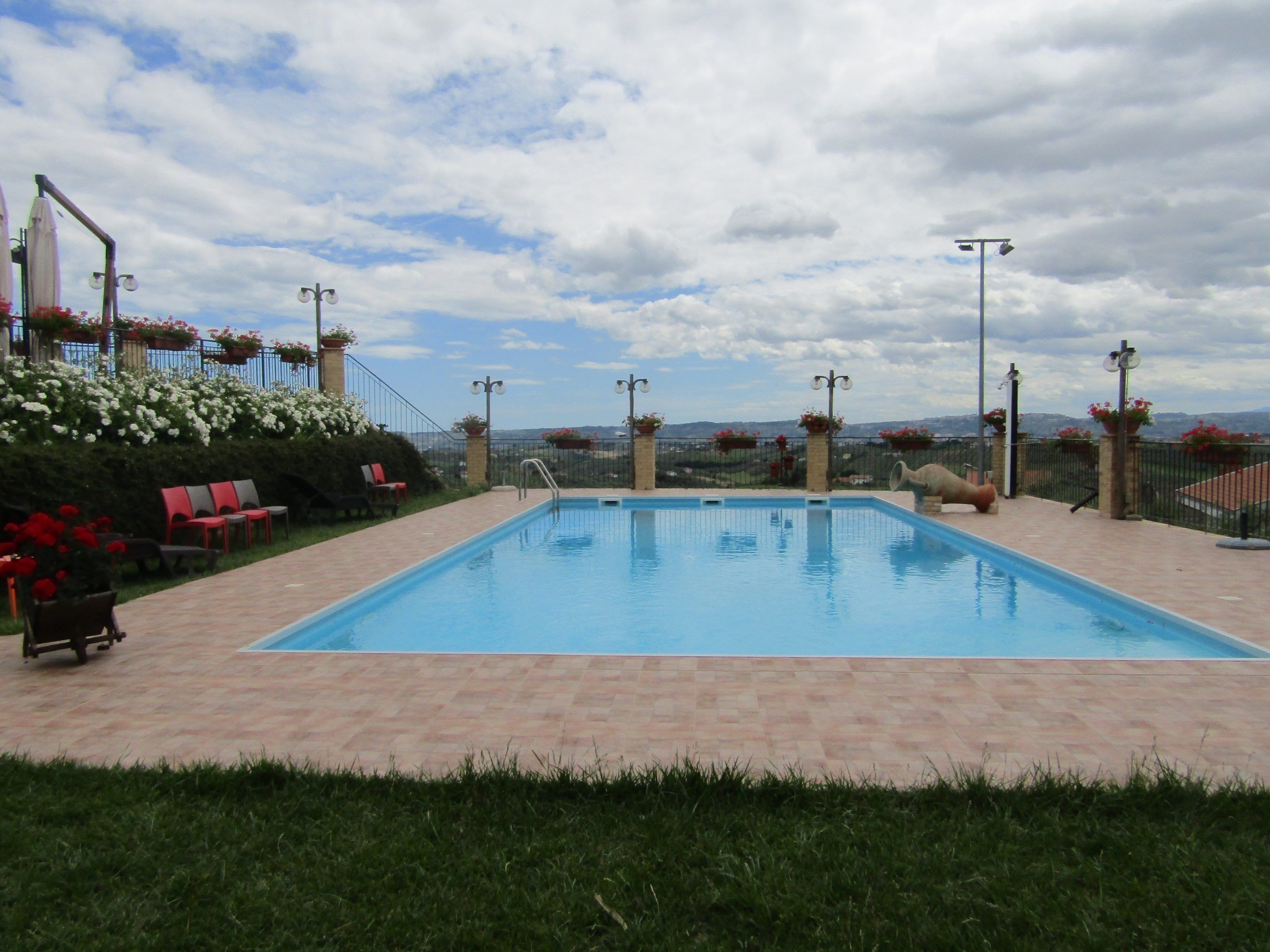 Una piscina rettangolare con bordo piastrellato, circondata da un basso muro in mattoni e una recinzione metallica. Lungo la recinzione si trovano sedie e piante in vaso.