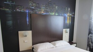 A hotel room with a large bed, two bedside tables, a nightstand, and a cityscape mural on the wall.