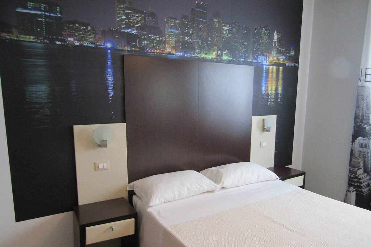 A hotel room with a large bed, two bedside tables, a nightstand, and a cityscape mural on the wall.