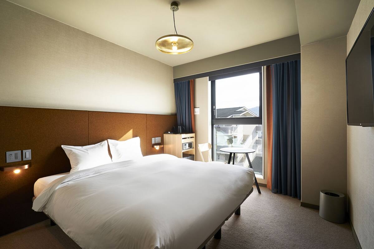 A hotel room with a large bed, a small table, a chair, a TV, and a window with curtains.