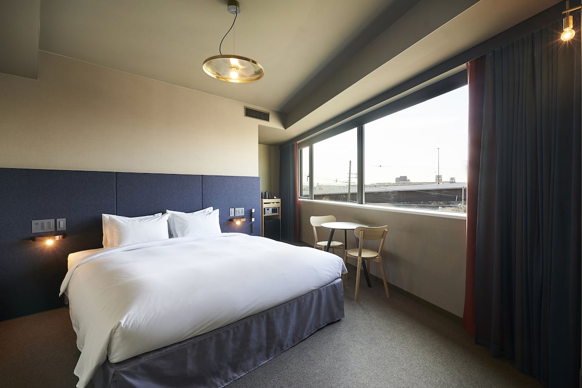 A hotel room with a large bed, a small table, and a window with a view of the city.