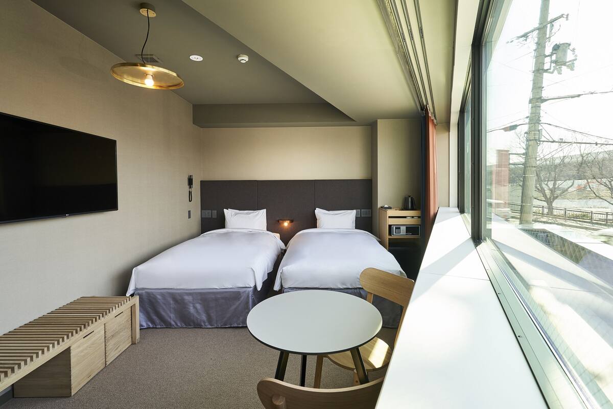 A hotel room with two beds, a small table, a chair, a TV, and a window with a view of the outside.