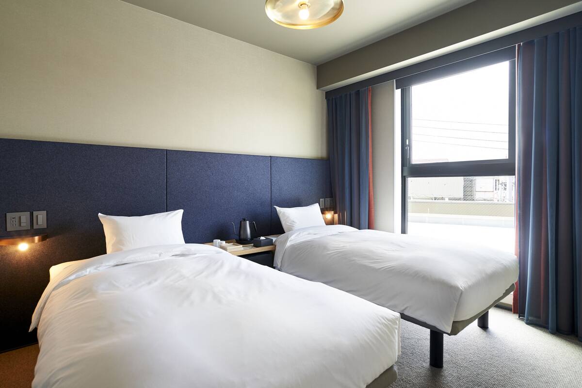 A hotel room with two beds, a headboard, bedside tables, a window with curtains, and a view of the outside.
