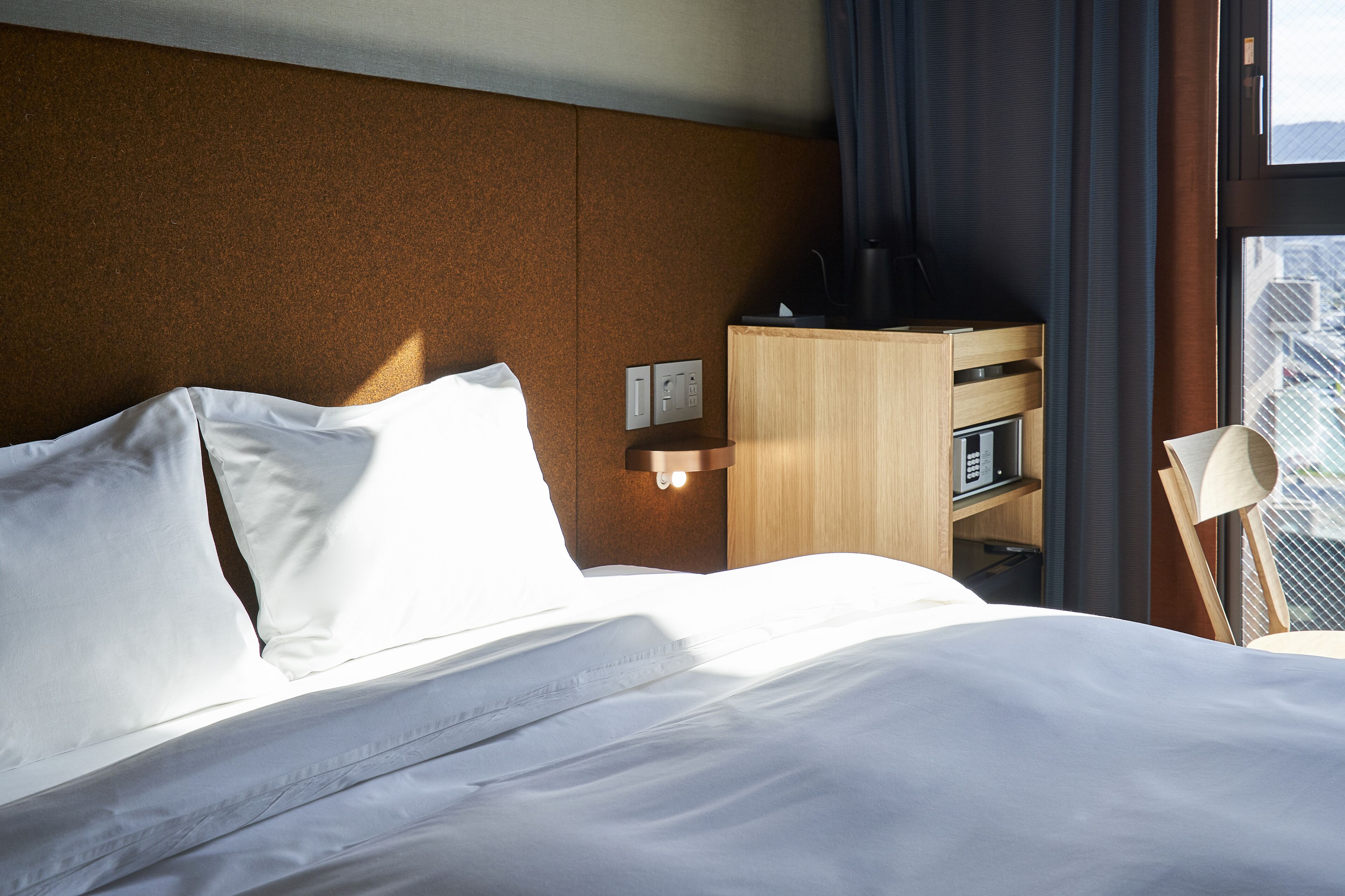 A hotel room with a bed, a wooden nightstand, a lamp, and a window with curtains.