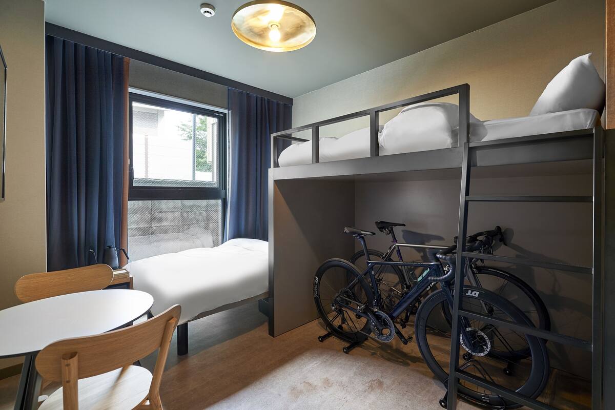 A compact hotel room with bunk beds, a bicycle rack, a small table, and a window with curtains.