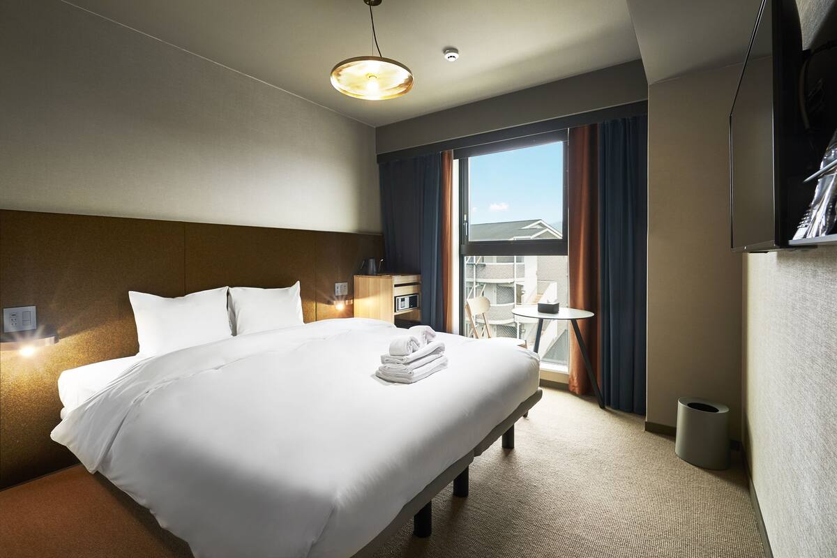 A hotel room with a large bed, a bedside table, a lamp, a window with curtains, and a view of the outside.
