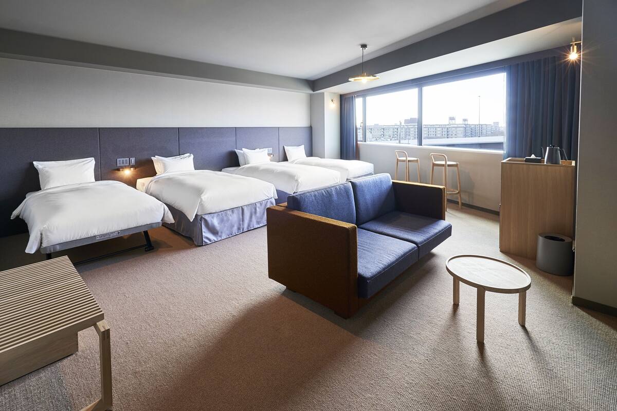 A hotel room with three beds, a sofa, a small table, and a window with a city view.