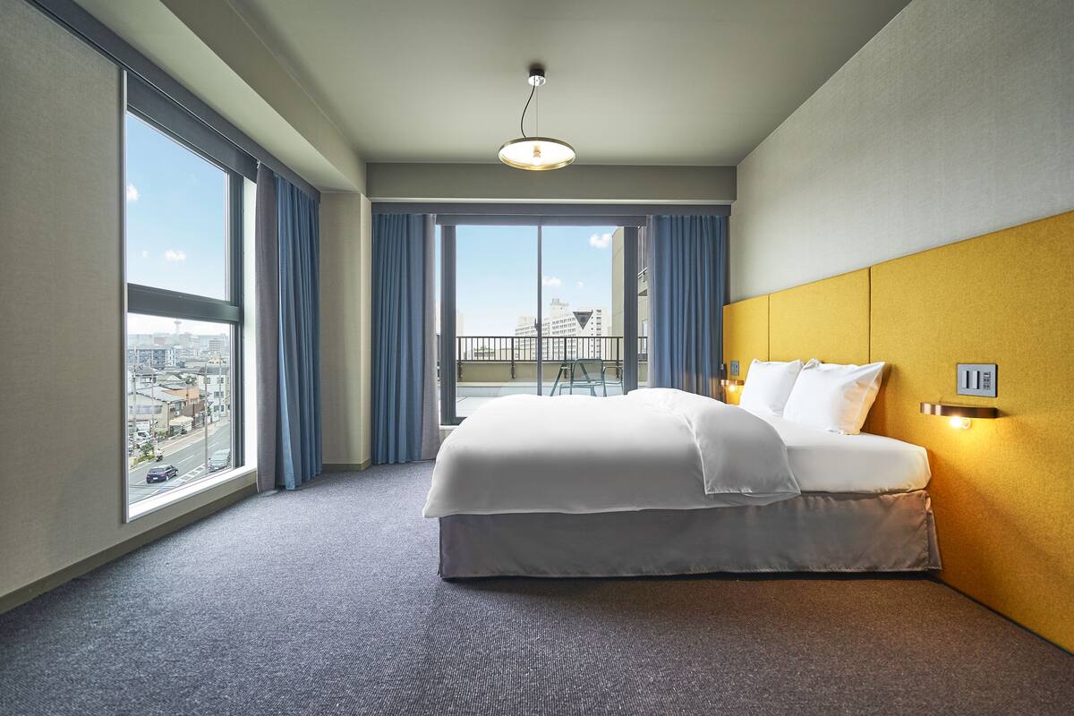 A hotel room with a large bed, a view of the city through floor-to-ceiling windows, and a balcony with a railing.