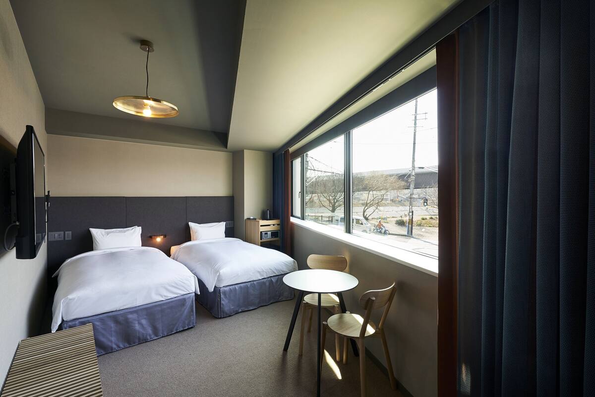 A hotel room with two beds, a small table, a chair, a TV, and a window with a view of the outside.