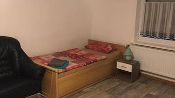 Una habitación pequeña con una cama de madera, una silla negra, una mesita y una ventana con cortinas.