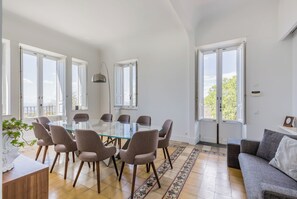 Uma sala de jantar com mesa e cadeiras de vidro, janelas amplas e um tapete estampado.