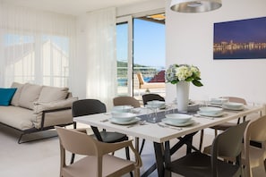 Uma área de refeições moderna com uma mesa branca posta para quatro pessoas, rodeada por cadeiras bege. Há um vaso com flores na mesa e uma pintura de paisagem urbana na parede.