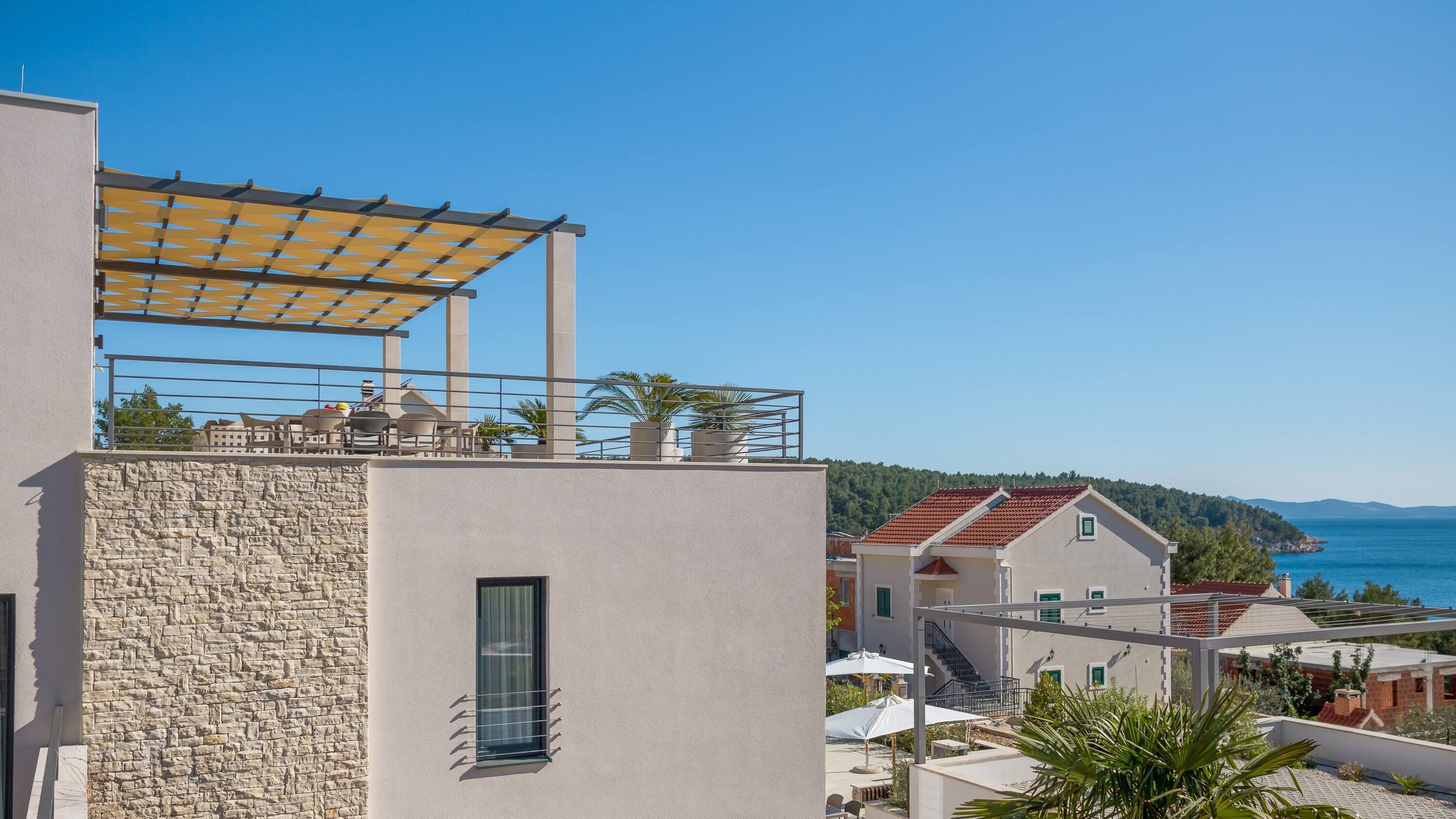 Un edificio moderno con facciata in pietra, una terrazza sul tetto con posti a sedere all'aperto e vista sul mare e le colline sullo sfondo.