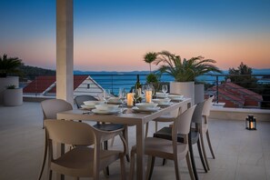 Ett matbord dukat för sex personer med tallrikar, glas och ljus på en terrass med utsikt över havet och bergen.