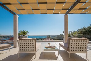 Terraço com duas cadeiras brancas, uma mesa pequena e uma bandeja com um copo e um recipiente. O terraço oferece uma vista panorâmica do mar e das colinas.