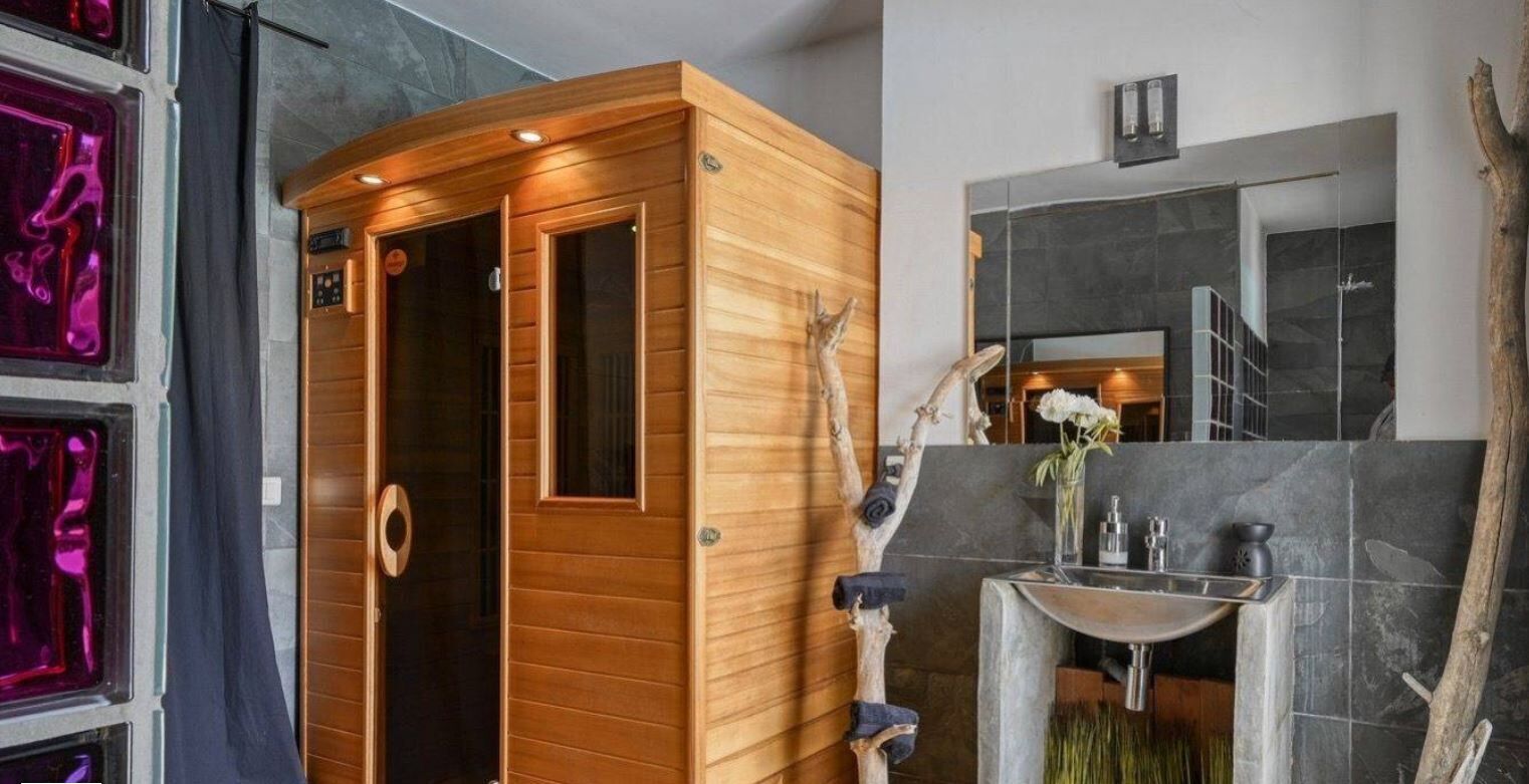 A sauna with a wooden exterior and a glass door, a stone-tiled wall, and a modern bathroom with a sink and mirror.