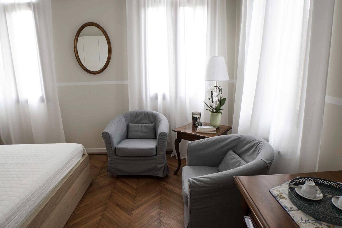 A hotel room with a bed, two grey armchairs, a small table with a lamp, and a mirror on the wall.