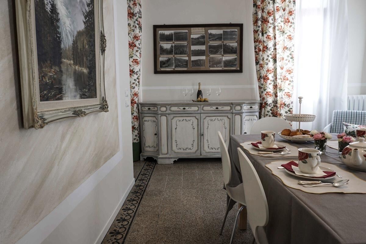 A dining area with a white cabinet, floral curtains, and a framed landscape painting.