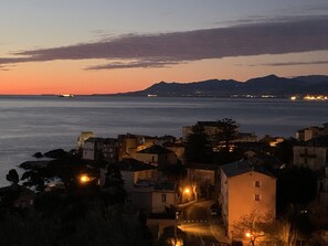 Un pueblo costero al atardecer con edificios a lo largo del malecón, un cielo despejado y montañas lejanas.