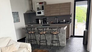 A modern kitchen with a built-in microwave, a coffee maker, and a sink. There are three bar stools and a sofa with a cushion. The floor is covered with dark tiles.