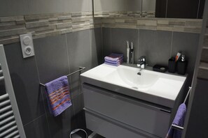 Un baño moderno con un lavamanos blanco, azulejos grises y un perchero con una toalla morada.