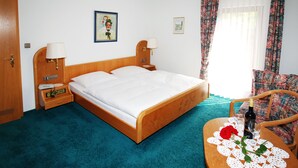 A hotel room with a wooden bed, two bedside tables with lamps, a small table with a bottle and glasses, a chair, and a window with curtains.