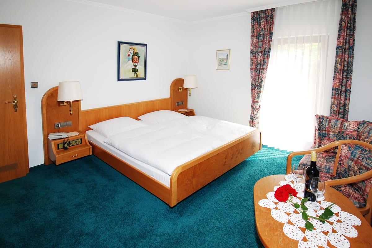 Habitación de hotel con cama de madera, dos mesitas de noche con lámparas, una mesita pequeña con una botella y vasos, una silla y una ventana con cortinas.