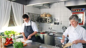 A professional kitchen with chefs preparing food, stainless steel appliances, and a variety of fresh vegetables on the counter.